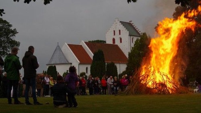 Folk samles omkring et stort bål nær en hvid kirke, hvor en folkemængde betragter flammerne.