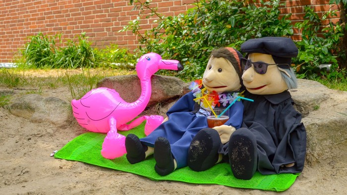 Zwei ausgestopfte Puppen sitzen neben einem rosa Flamingo auf einer grünen Matte im Freien.
