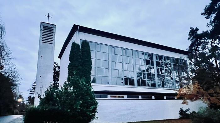 Moderne Kirchengebäude mit großen Glasfenstern und einem hohen Glockenturm neben üppigem Grün.