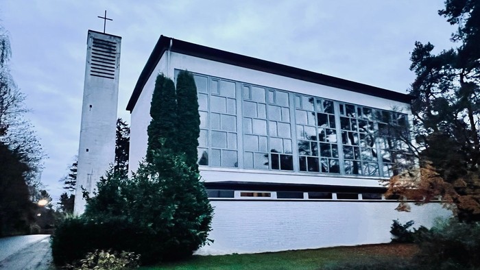 Moderne Kirchengebäude mit großen Glasfenstern und einem hohen Glockenturm neben üppigem Grün.
