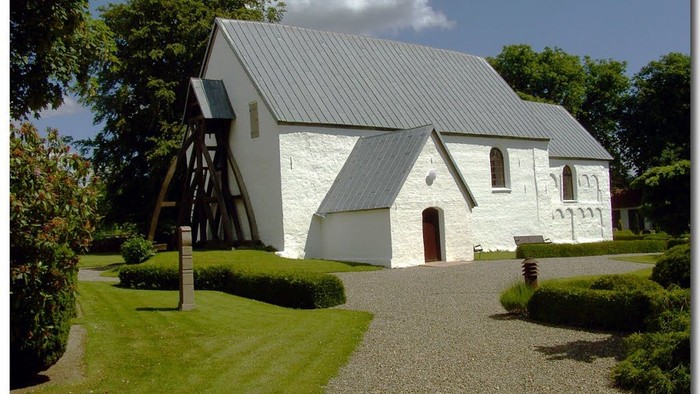 Lille hvid kirke med gråt tag, omgivet af grønne omgivelser.