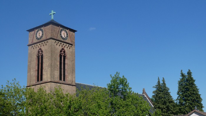 Ein hoher Backsteinkirchturm mit Uhren und einem Kreuz vor einem klaren blauen Himmel.