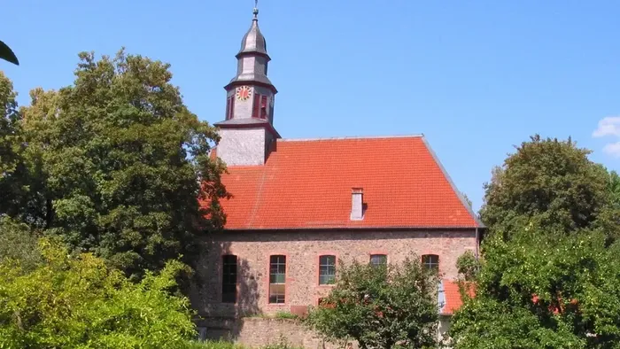 Idyllische Steinkirche mit rotem Dach und Glockenturm, umgeben von üppigem Grün unter klarem Himmel.