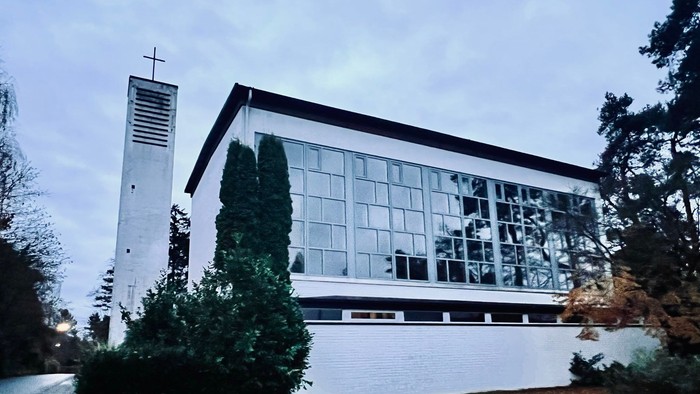 Moderne Kirchengebäude mit großen Glasfenstern und einem hohen Glockenturm neben üppigem Grün.
