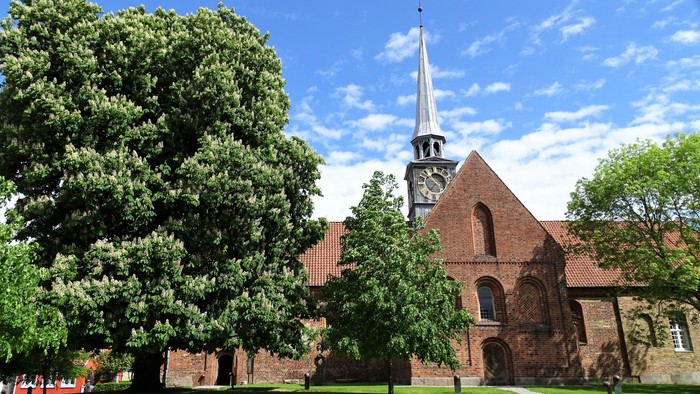 En rød murstenskirke med tårn og klokke, omgivet af grønne træer og en græsplæne