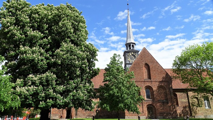En rød murstenskirke med tårn og klokke, omgivet af grønne træer og en græsplæne