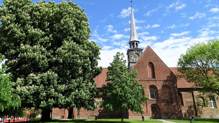 En rød murstenskirke med tårn og klokke, omgivet af grønne træer og en græsplæne