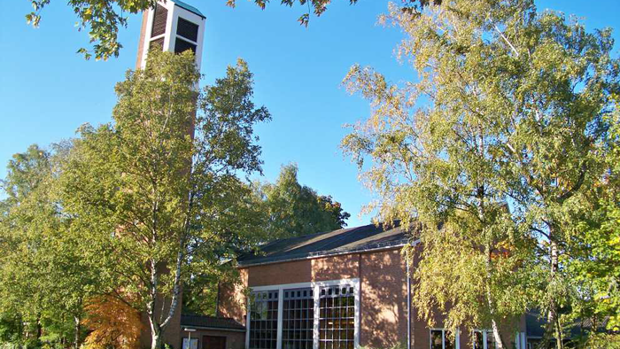 Eine malerische Backsteinkirche mit einem hohen Glockenturm, umgeben von üppigen Herbstbäumen unter einem klaren blauen Himmel.