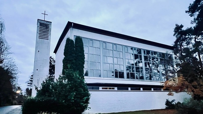 Moderne Kirchengebäude mit großen Glasfenstern und einem hohen Glockenturm neben üppigem Grün.
