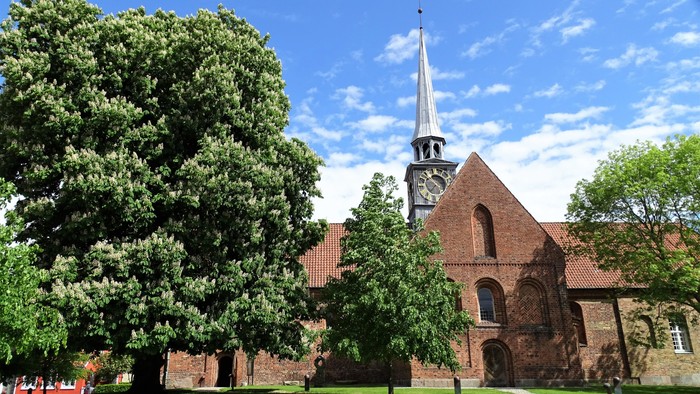En rød murstenskirke med tårn og klokke, omgivet af grønne træer og en græsplæne