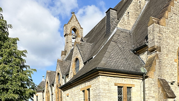 Historische Steinkirche mit Spitzbögen, steilem Dach und angrenzenden Bäumen unter einem teilweise bewölkten Himmel.