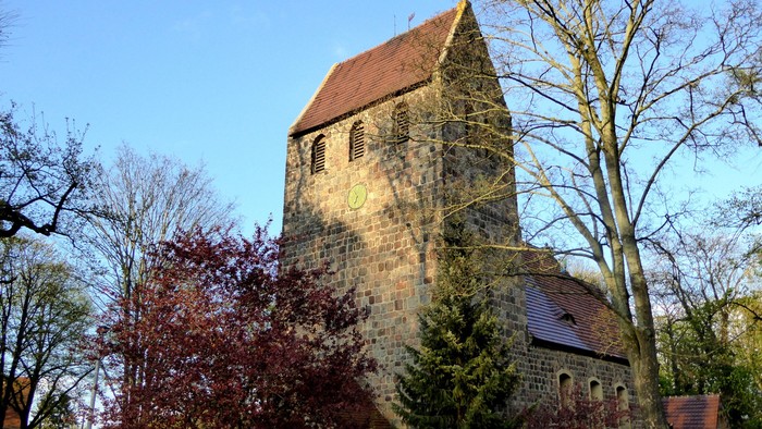 Alte Kirche mit hohem Turm und rotem Dach, umgeben von Bäumen und Büschen