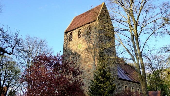 Alte Kirche mit hohem Turm und rotem Dach, umgeben von Bäumen und Büschen
