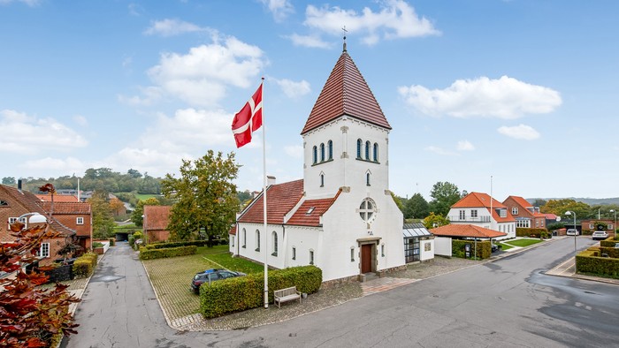 En lille hvid kirke med rødt tag og en dansk flag på en grøn plads i et landsbyområde