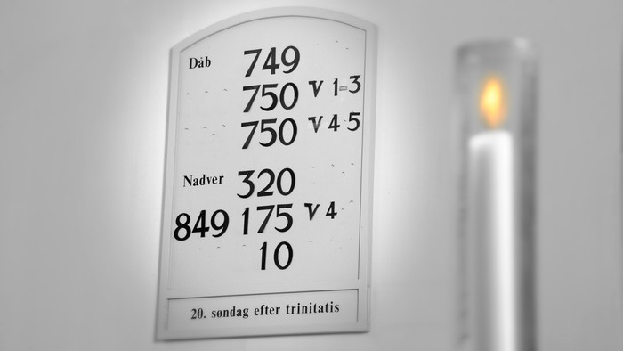 Et tændt lys ved siden af en kirkegudstjeneste-salmebog med nummererede læsninger og datoer på dansk.