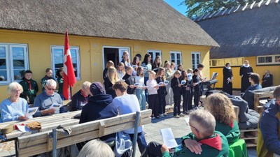 Udendørs korøvelse med folk, der synger nær en stråtagshytte.