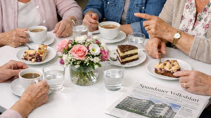 Eine Gruppe genießt Tee, Kuchen und Gespräche an einem Tisch mit Blumen und einer Zeitung.