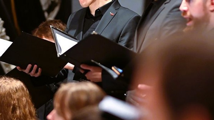 Ein Chor singt aus Notenheften, während das Publikum zuhört.