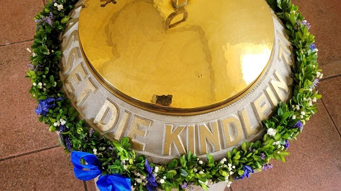 Verzierter Taufstein mit Blumenkranz und Band, beschriftet mit *„De Kindlamp“* auf dem Sockel.