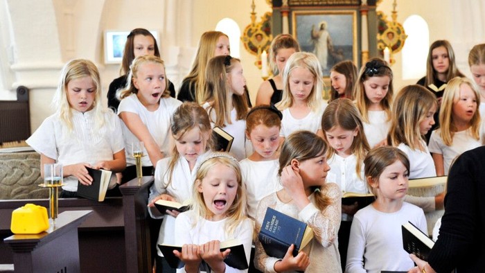 En gruppe børn synger i en kirke under ledelse af en kvinde.