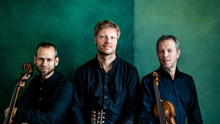 Drei Musiker sitzen zusammen und halten ein Cello, eine Gitarre und eine Geige vor einem grünen Hintergrund.