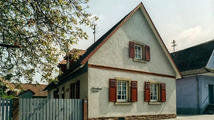 Das Bild zeigt ein charmantes, kleines Haus mit rotem Dach und weißen Fensterläden an einer Kopfsteinpflasterstraße.