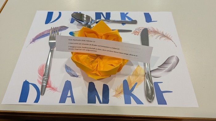 Eine akkurat gedeckte Tischdecke mit einer gefalteten Serviette, Besteck und einem Zettel mit der Aufschrift „Danke“ auf einer dekorativen Platzdecke.
