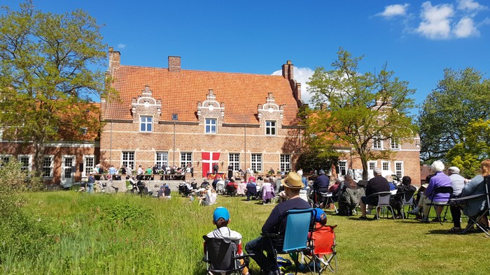 En gruppe af mennesker sidder på stolene udenfor et stort, rødt teglstenshus med en blå flag.
