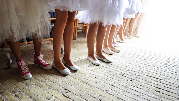 En række mennesker i balletsko og balletnederdele, hvor én har afslappede sneakers på, står i et lokale.