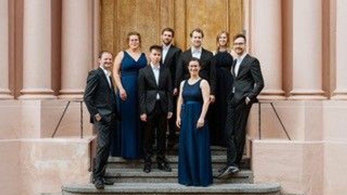 ine Gruppe von acht Personen steht auf den Stufen vor einem Gebäude mit hohen Säulen und einer großen Holztür. Die Mitglieder sind festlich gekleidet: Die Männer tragen dunkle Anzüge, die Frauen elegante Kleider, teils in dunklen, teils in kräftigen Farbt