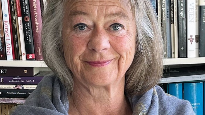 En smilende kvinde med gråt hår står foran en bogreol fyldt med forskellige bøger.