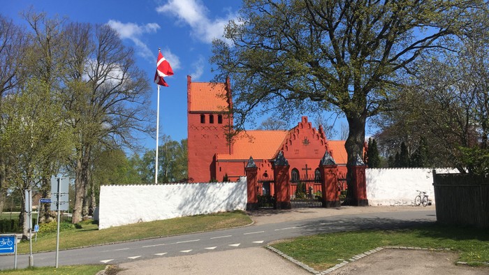 Livligt rødt slotlignende bygning med en flagstang, der viser et skandinavisk flag, omgivet af træer.