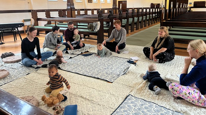 En gruppe mennesker og børn samles indendørs på måtter i en rummelig sal med træbænke.