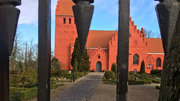 En slående rødstenskirke set gennem en metalramme, indrammet af nøgne træer og en brostensbelagt sti.