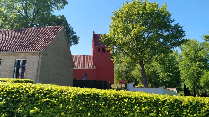 Hyggelig stenkirke med rødt spir omgivet af frodig grønne omgivelser under klar blå himmel.