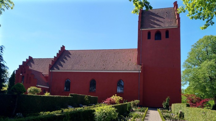 Livligt rødt kirkebyggeri med frodige havegange og veltrimmede hække under klar blå himmel.