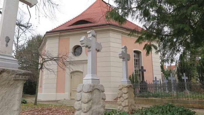 Friedliche Kirche mit Kreuzen auf steinernen Sockeln in einem ruhigen Friedhof, umgeben von Bäumen.