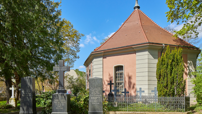Ein kleiner rosa und weißer Glockenturm mit spitzem Dach in einem Friedhof, umgeben von Bäumen und Grabsteinen.