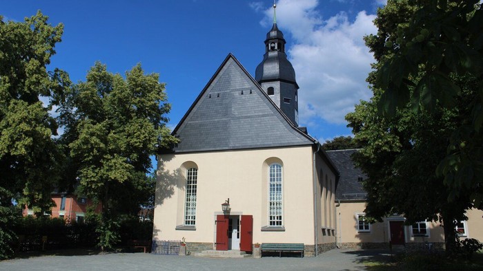 Idyllische Kirche mit steilem Satteldach, umgeben von üppigen grünen Bäumen unter einem strahlend blauen Himmel.