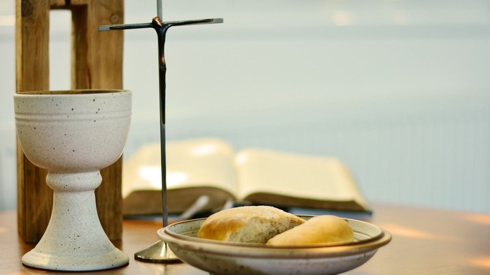 Ein Kelch, ein Kreuz und ein offenes Buch liegen auf einem Tisch neben einem Teller mit Brot.
