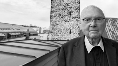 Ein älterer Mann in formeller Kleidung steht auf einem Dach und blickt auf Industriegebäude hinab.