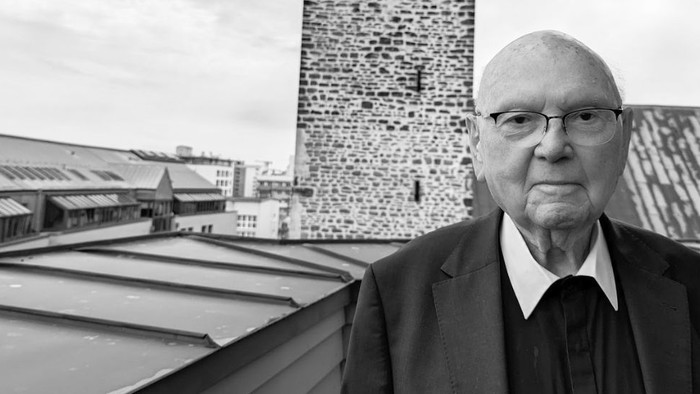 Ein älterer Mann in formeller Kleidung steht auf einem Dach und blickt auf Industriegebäude hinab.