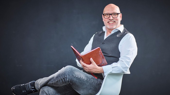 Mann mit Brille sitzt lässig und hält ein rotes Buch.