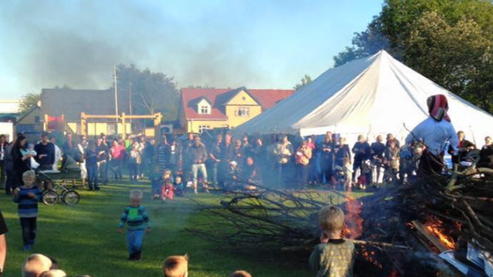 En folkemængde ser en stor bål på en grøn plads med telt og huse i baggrunden