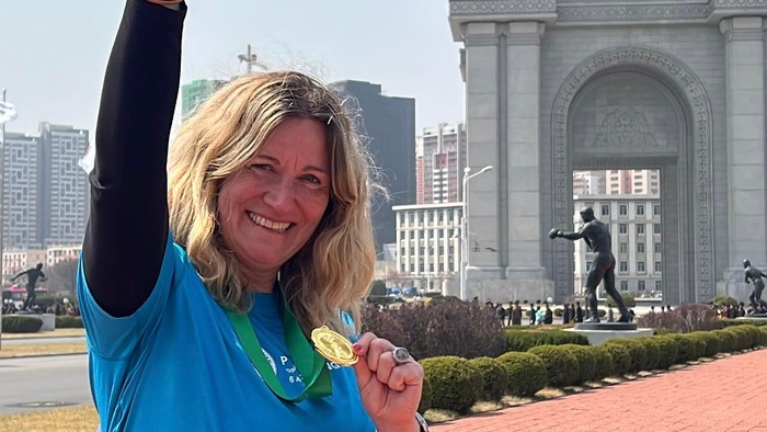 Glædestrålende løber viser stolt medalje og startnummer frem foran et stort monument.