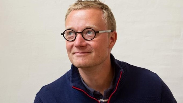 En smilende mand med briller iført en mørkeblå sweater med rød kant foran en ensfarvet væg.