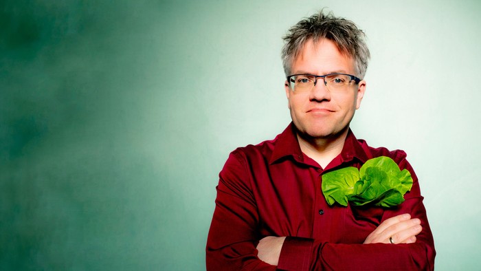 Ein Mann in einem roten Hemd hält eine belaubte grüne Pflanze mit verschränkten Armen vor der Brust.