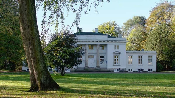 Ein großes weißes Haus mit Säulen und vielen Fenstern, umgeben von Bäumen und einem grünen Rasen.