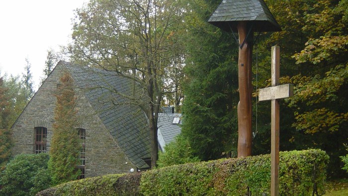Idyllische Steinkapelle, umgeben von Grün, mit einem hölzernen Kreuz und einem kleinen Pavillon in der Nähe.