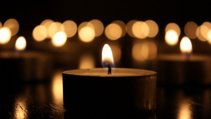 Single lit candle in dark room with many other candles in background.